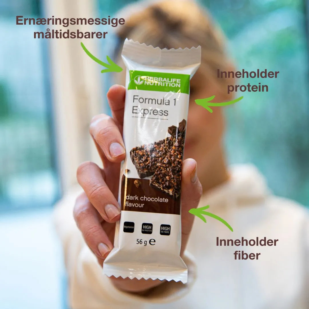 En person holder en Herbalife Formula 1 mellombar med mørk sjokolade. På bildet står tekstene “Ernæringsmessige måltidsbarer”, “Inneholder protein” og “Inneholder fiber” med piler som peker mot mellombaren.