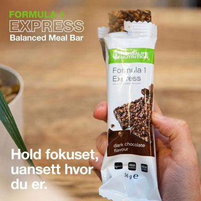 En person holder en åpnet Herbalife expressbar mellombar med mørk sjokolade. På bildet står tekstene "Formula 1 Express Balanced Meal Bar" og “Hold fokuset, uansett hvor du er.”.