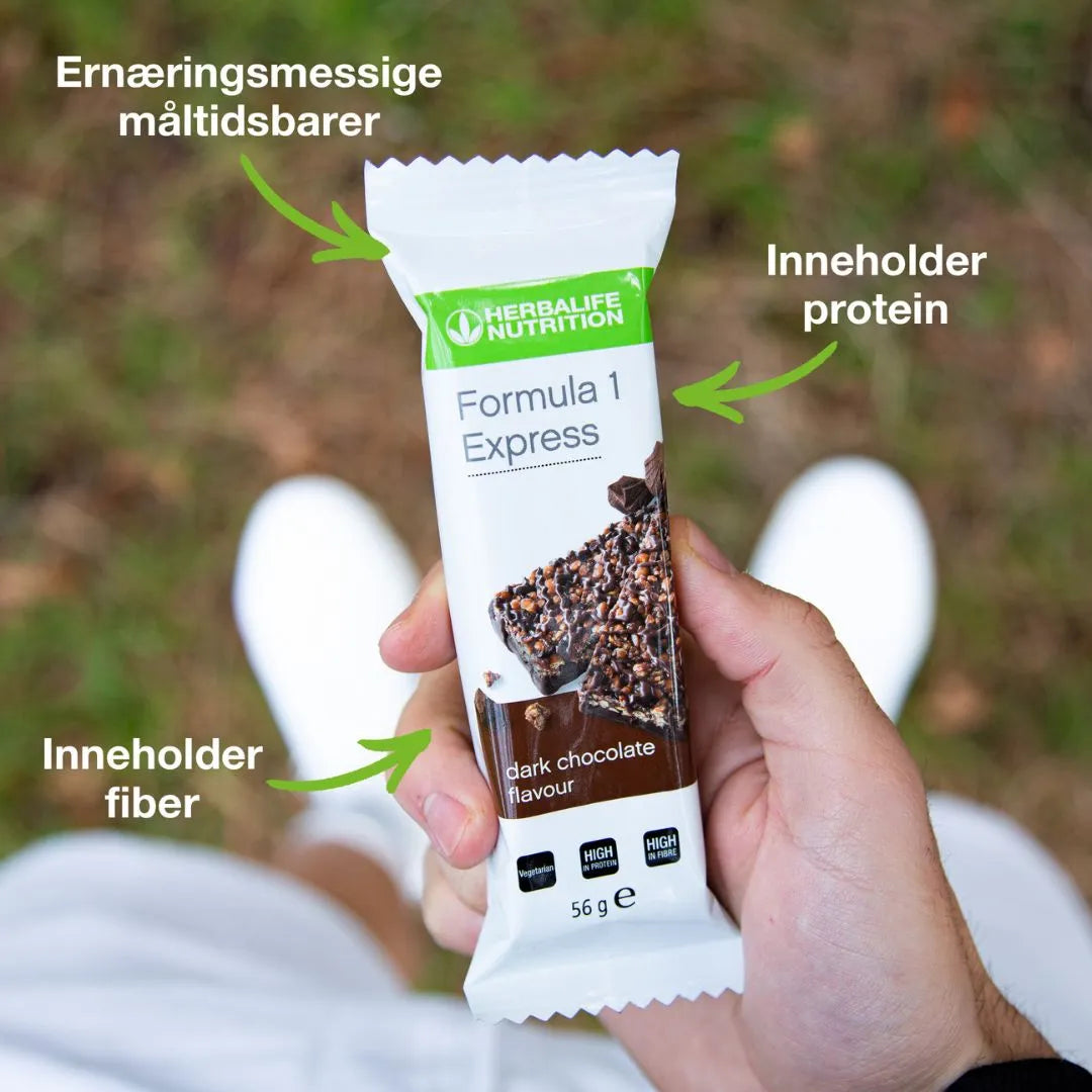 En person holder en Herbalife Formula 1 mellombar med mørk sjokolade. På bildet står tekstene “Ernæringsmessige måltidsbarer”, “Inneholder protein” og “Inneholder fiber” med piler som peker mot mellombaren.