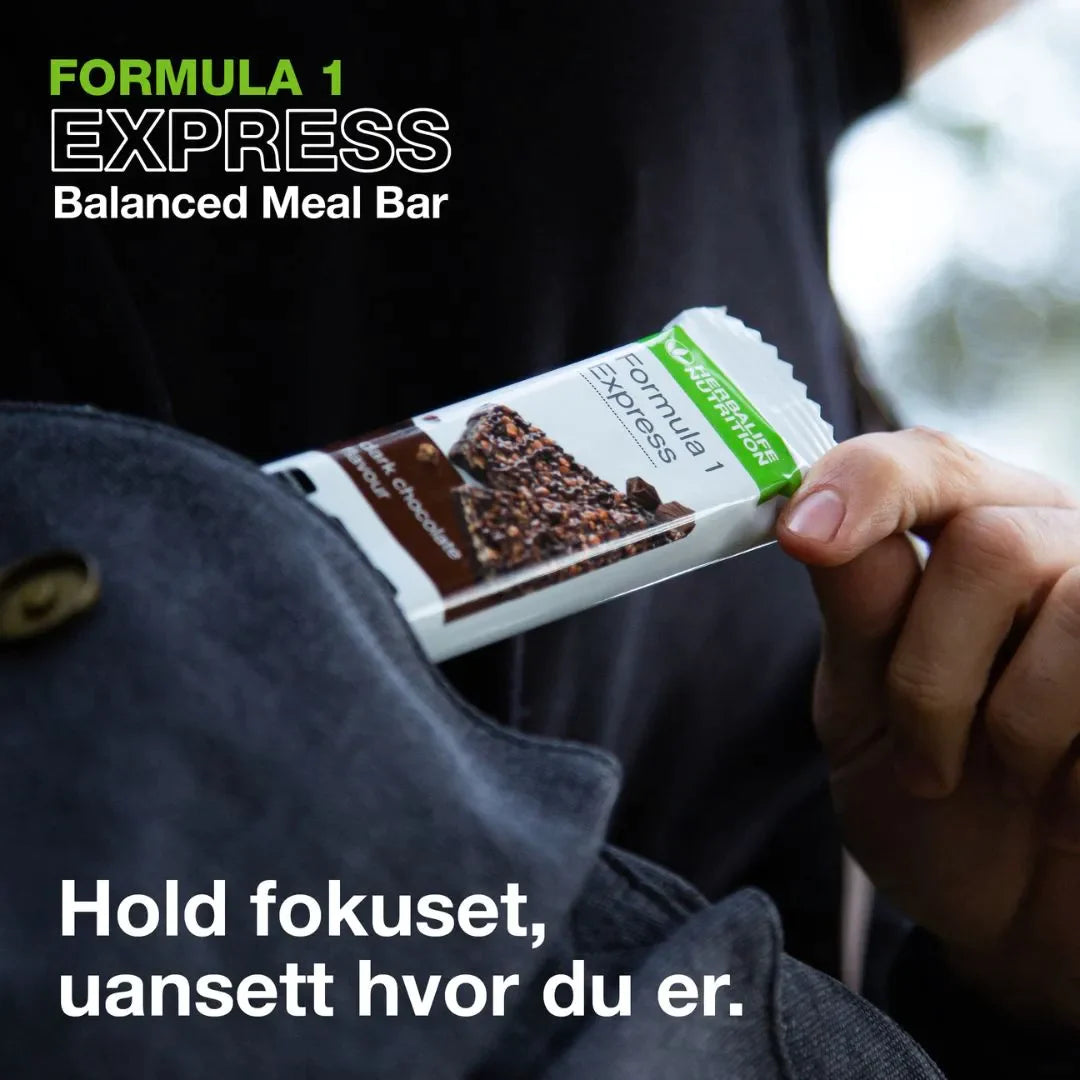 En person i en mørkeblå jakke tar en Herbalife Formula 1 mellombar med mørk sjokolade opp av jakkelomma. På bildet står teksten "Formula 1 Express Balanced Meal Bar".