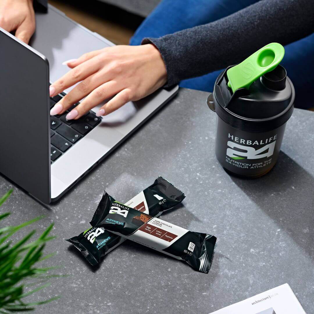 To Herbalife H24 Achieve proteinbarer med mørk sjokolade ligger på et bor hvor det sitter en kvinne og jobber på en PC. Ved siden av står også en shaker med Herbalife H24-logo. 