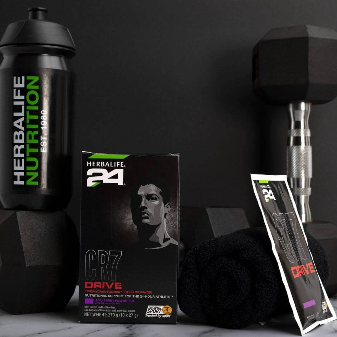 En pakke Herbalife H24 CR7 Drive porsjonspose og en løs porsjonspose står sammen med en sportsdrikkeflaske med Herbalife Nutrition-logo, noen dumbbells og et svart håndkle. 