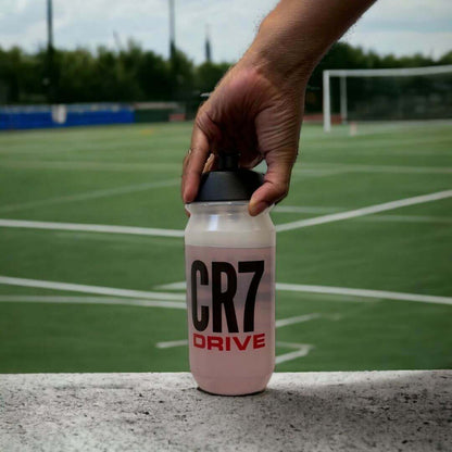 En hånd strekker seg etter en drikkeflaske med Herbalife H24 CR7 Drive sportsdrikk som står ved en fotballbane. Drikkeflaska har teksten CR7 Drive.