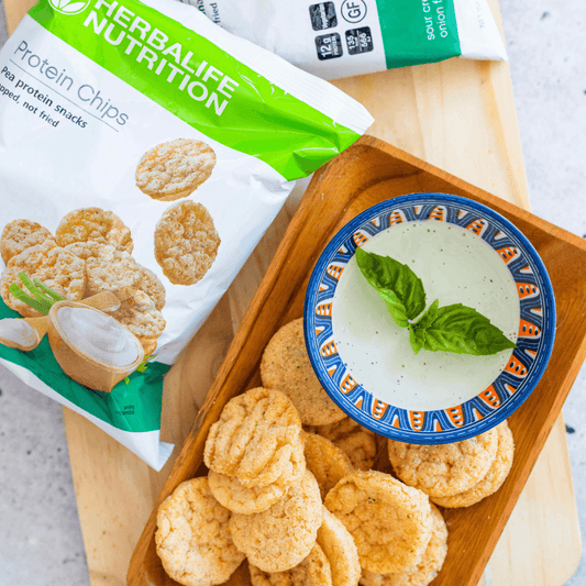 En porsjonspose Herbalife Protein Chips proteinrik snacks sourcream & onion-smak ligger på ei lys trefjøl sammen med et fat med proteinchips og ei skål med dip.