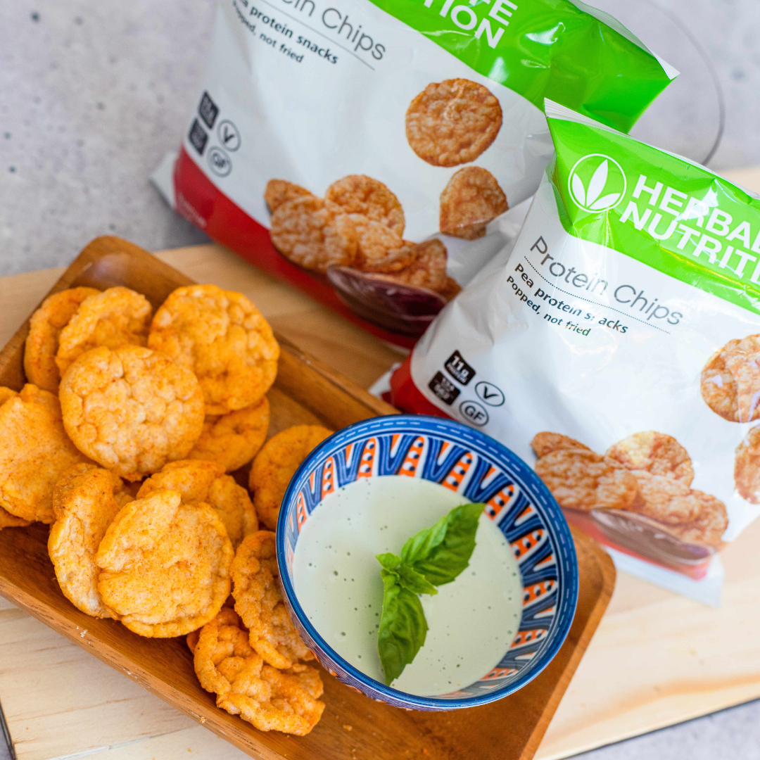 To porsjonsposer Herbalife Protein Chips proteinrik snacks. En pose med sourcream & onion-smak og en pose med barbeque-smak, står på en lys trefjøl sammen med et fat med proteinchips og en skål med dip.