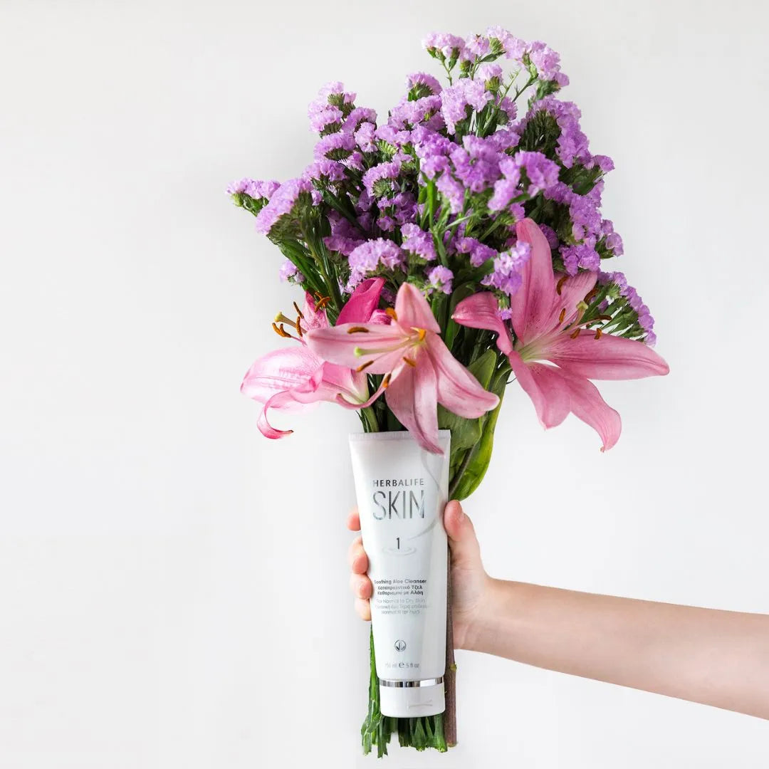 En hånd holder en tube Herbalife SKIN Soothing Aloe Cleanser foran en blomsterbukett med rosa og lilla blomster. 