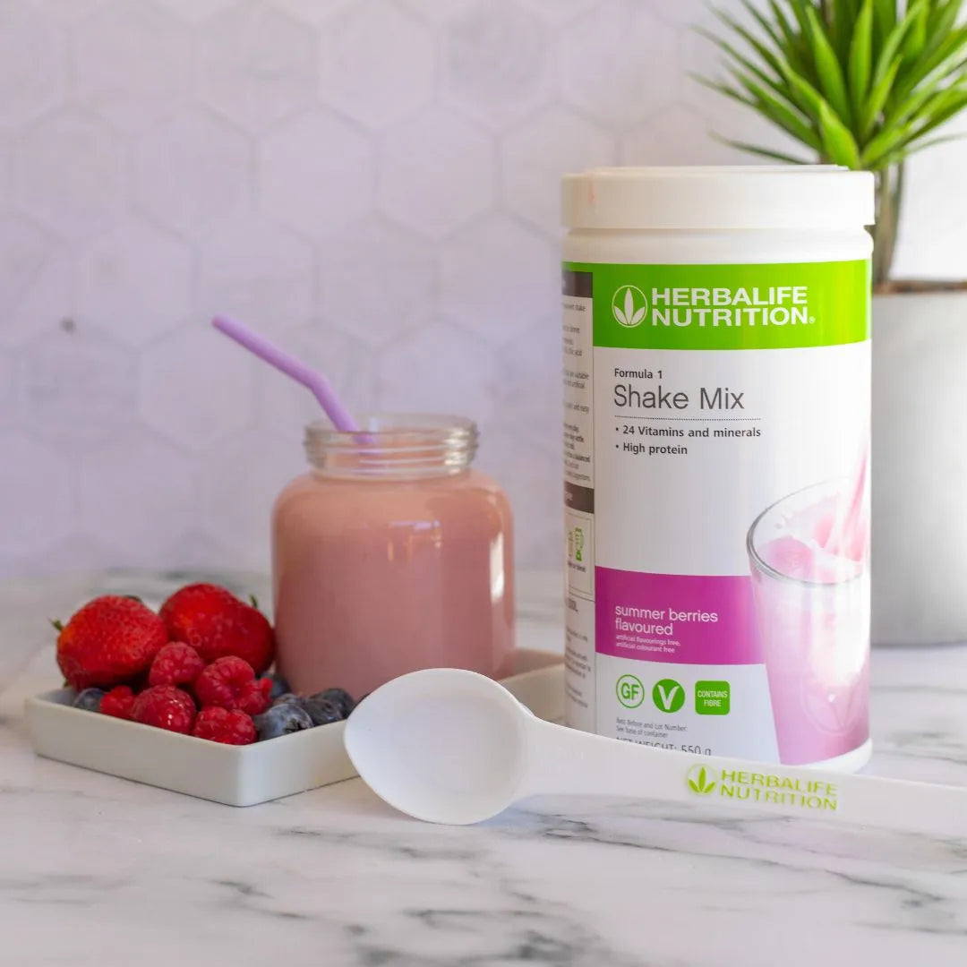 En boks Herbalife Formula 1 shakemix med Summer Berries-smak sammen med en rosa sommerbær-smoothie i et glasskrus med lilla sugerør. På bordet ligger røde bær; jordbær, bringebær og blåbær, samt en hvit måleskje med teksten Herbalife Nutrition.