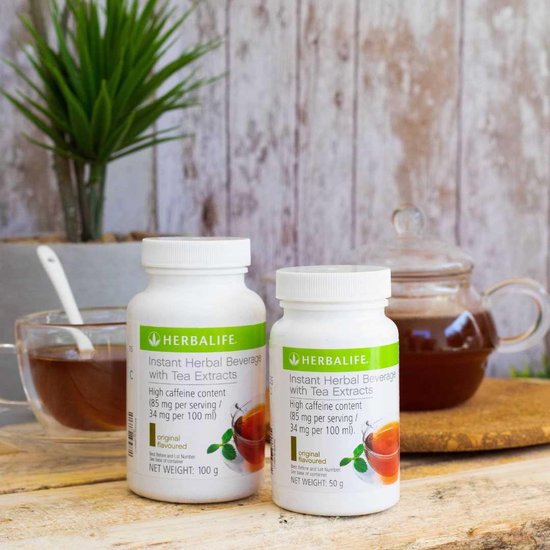 2 bokser Herbalife instant herbal beverage urtete med originalsmak i to ulike størrelser, en glasskopp og ei glass-tekanne med te på en trebenk. 