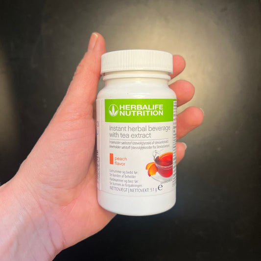 En boks Herbalife Instant Peach urtekonsentrat te med ferskensmak, 51 gram, i hånda.