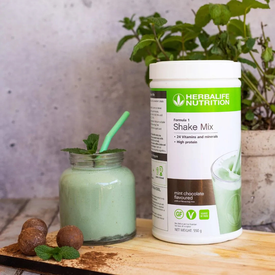 En boks Herbalife Formula 1 shakemix med Mint chocolate-smak sammen med et glasskrus med mintsjokolade-shake og et mintgrønt sugerør. Tre sjokoladekuler strødd med kakao og et mintblad ligger foran glasset. I bakgrunnen ses en mynteplante.