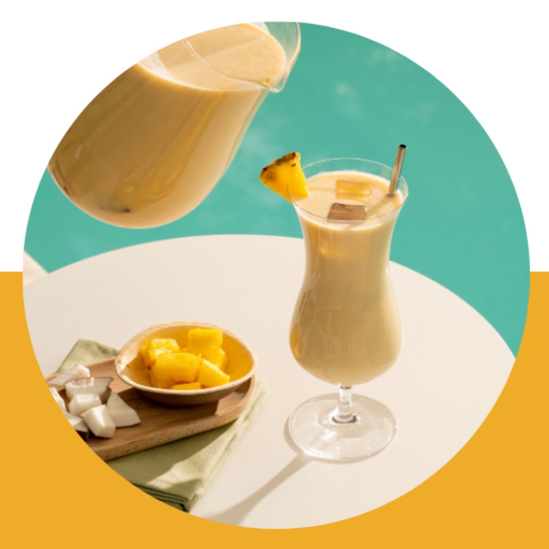 En smoothie i et høyt drinkglass, pyntet med en skive ananas. Et brett med kokos og ananas står ved siden av og det er ei mugge med smoothie i. 