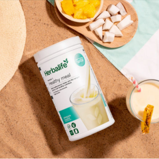 En boks Herbalife-shake med pineapple coconut-smakligger på ei strand ved siden av en strandhatt, en smoothie og et brett med ananas og kokosnøtt på et håndkle. 