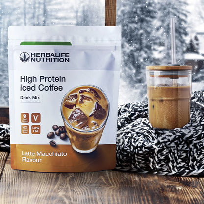 En pakke Herbalife High Protein Iced Coffee Drink Mix Latte Machiato-smak og et glasskrus med en protein-iskaffe med et skjerf rundt, i et vindu med vinterlandskap ute.