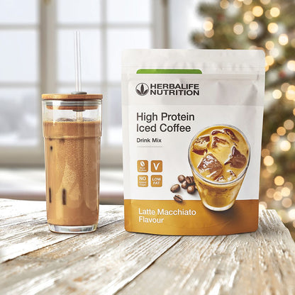 En pakke Herbalife High Protein Iced Coffee Drink Mix Latte Machiato-smak og et glasskrus med en protein-iskaffe på et bord og i bakgrunnen skimtes et juletre.