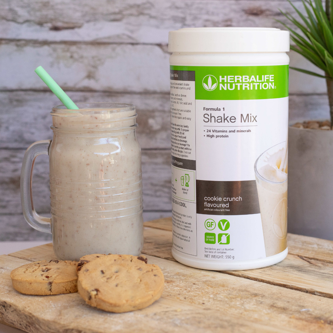 Herbalife Formula 1 shakemix med Cookie Crunch-smak og en Herbalife-shake laget som smoothie servert i et glassgrus. Foran ligger tre sjokoladecookies. 