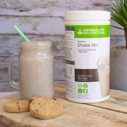 Herbalife Formula 1 shakemix med Cookie Crunch-smak og en Herbalife-shake laget som smoothie servert i et glassgrus. Foran ligger tre sjokoladecookies. 