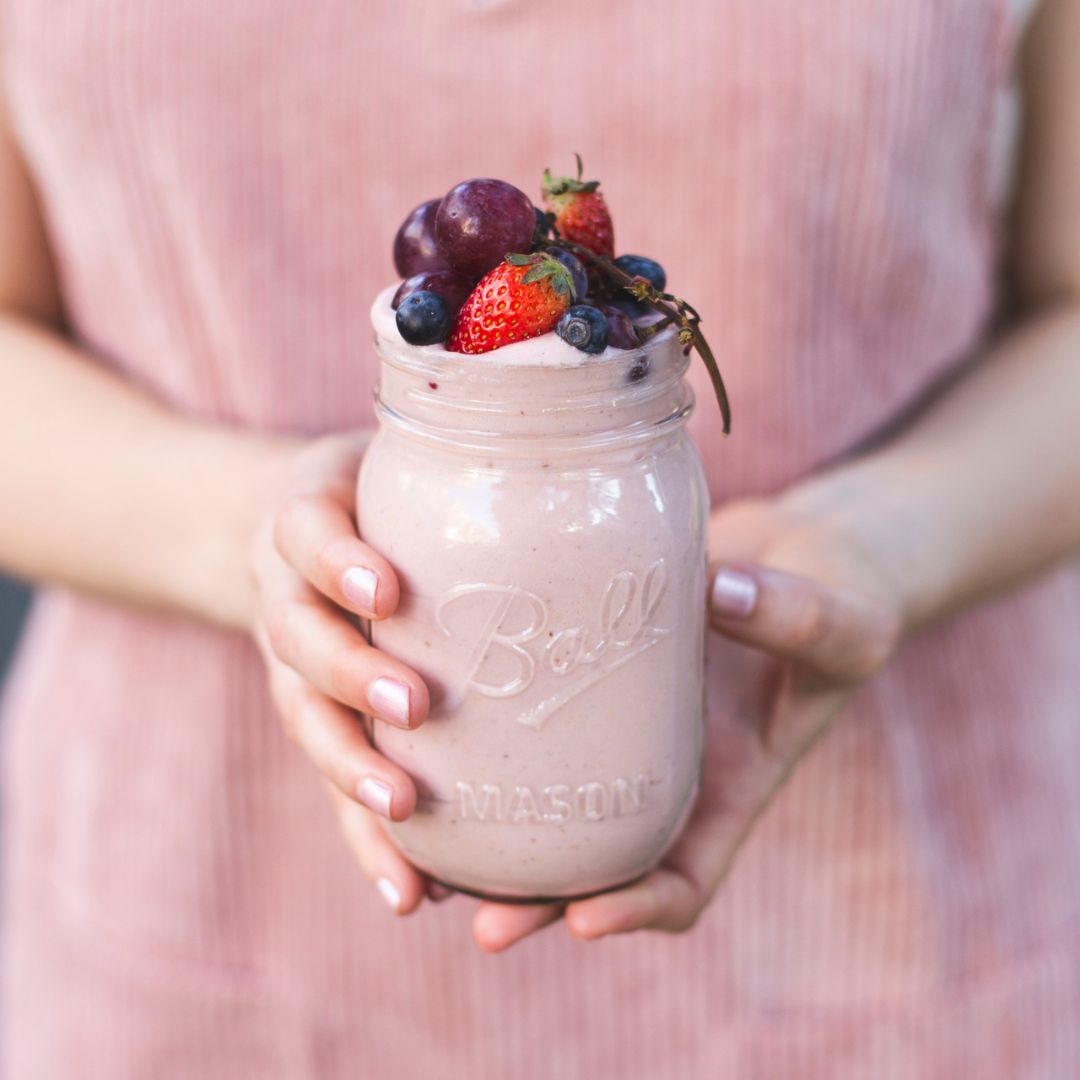 En kvinne holder et glasskrus med en rosa Herbalife-shake laget som en smoothie, toppet med ferske jordbær, røde druer og blåbær. 