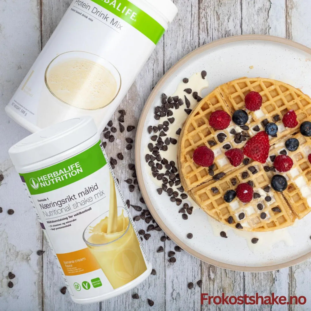 En boks Herbalife Formula 1 shakemix banansmak og en boks Proteindrinkmiks ligger på et bord sammen med et fat med en belgisk vaffel toppet med gresk yoghurt, bringebær, jordbær, blåbær og overstrødd med kakaonibs.