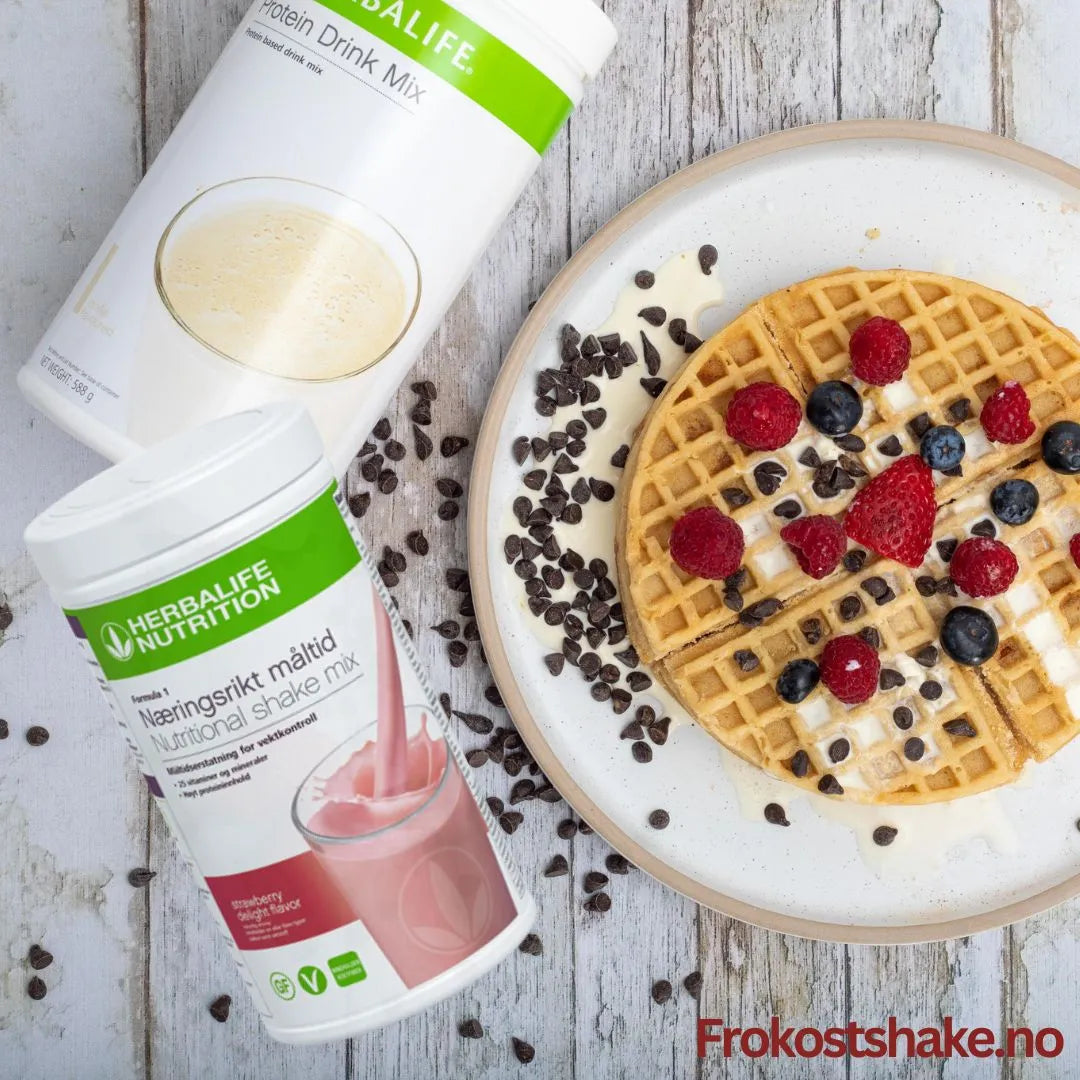 En boks Herbalife Formula 1 shakemix jordbærsmak og en boks Proteindrinkmiks ligger på et bord sammen med et fat med en belgisk vaffel toppet med gresk yoghurt, bringebær, jordbær, blåbær og overstrødd med kakaonibs.