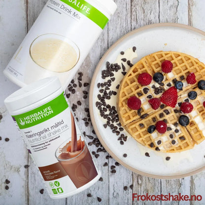 En boks Herbalife Formula 1 shakemix sjokoladesmak og en boks Proteindrinkmiks ligger på et bord sammen med et fat med en belgisk vaffel toppet med gresk yoghurt, bringebær, jordbær, blåbær og overstrødd med kakaonibs.