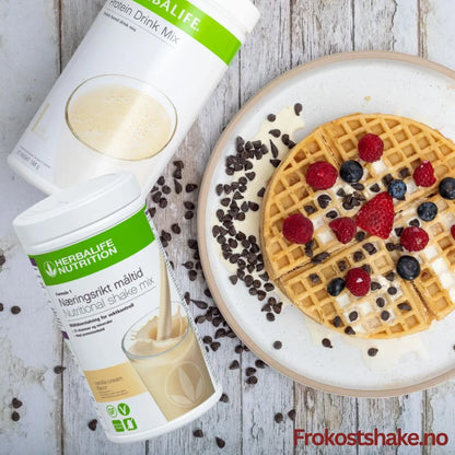 En boks Herbalife Formula 1 shakemix vaniljesmak og en boks Proteindrinkmiks ligger på et bord sammen med et fat med en belgisk vaffel toppet med gresk yoghurt, bringebær, jordbær, blåbær og overstrødd med kakaonibs.