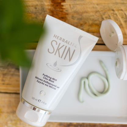 En tube Herbalife SKIN Purifying Mint Clay Mask ansiktsmaske ligger på et fat sammen med en åpnet tube og av produktet, en grønn leiremaske, er klemt ut.  