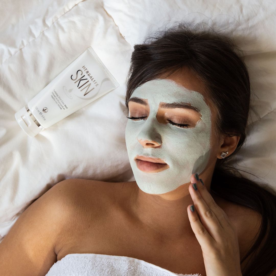 En tube Herbalife SKIN Purifying Mint Clay Mask grønn ansiktsmaske ligger ved siden av en kvinne som har grønn ansiktsmaske og ligger med et håndkle over seg i sengen.