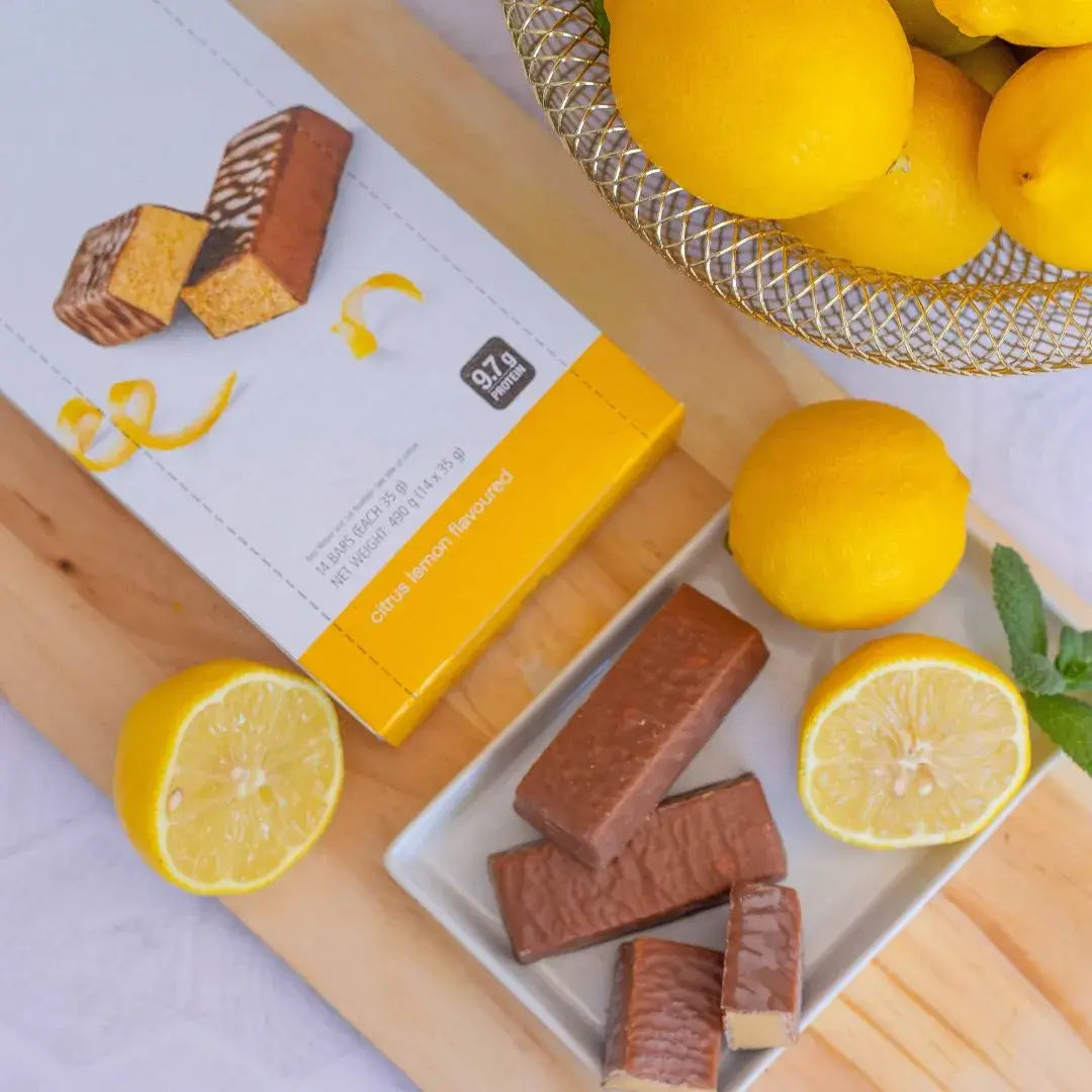 Sitron proteinbar fra Herbalife i eske og tre barer uten papir som ligger på et fat sammen med noen sitroner. 