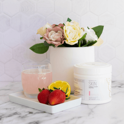 En boks Herbalife SKIN Collagen Booster står på et bord sammen med et glass drikkeklar kollagen, to jordbær og en halv sitron. Bak står en blomstervase. 