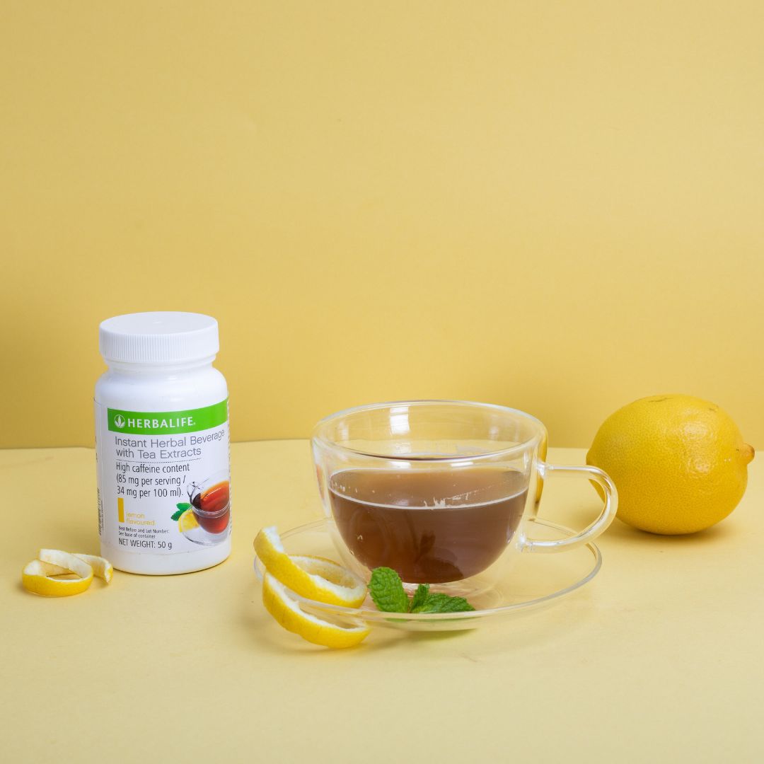 En glasskopp sitronte med sitronskall og mynteblad på skåla. En hel sitron. En boks Herbalife Instant Lemon urtekonsentrat te med sitronsmak, 51 gram.