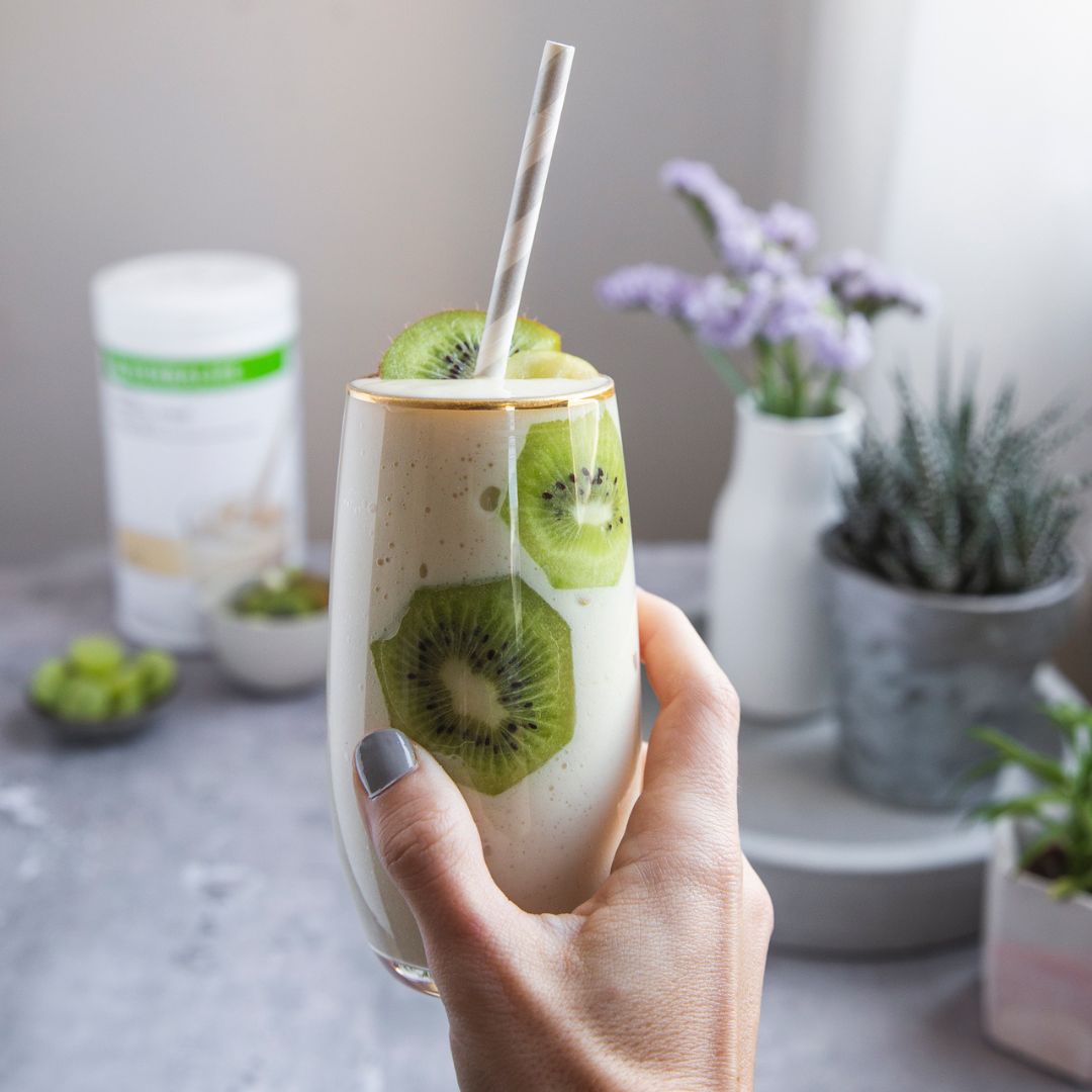 I forgrunnen holder en kvinnehånd en Herbalife-smoothie i et glass pyntet med skiver av kiwi på innsiden og på toppen. I den uklare bakgrunnen skimtes en boks med Herbalife Formula 1 shakemix vanilje, og to skåler med frukt.