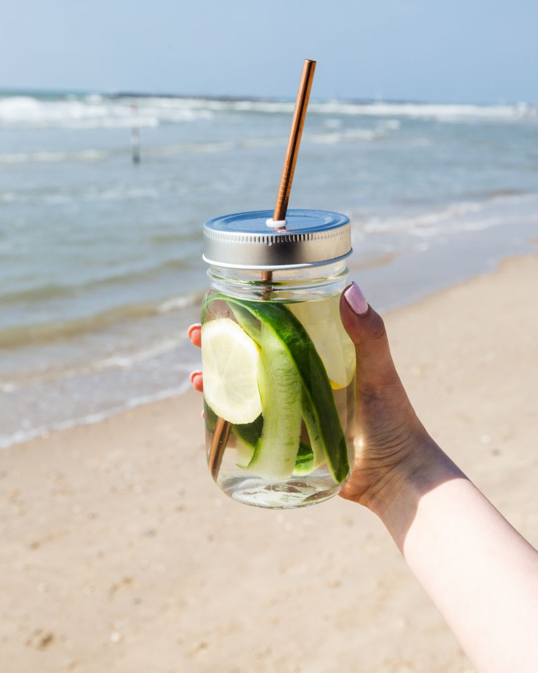 En hånd holder opp et krus med Herbalife aloeverajuice på ei strand. I glasset er det skivet sitron og agurk.