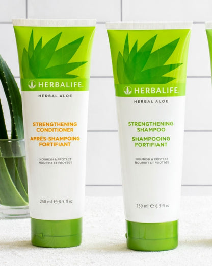 En flaske Herbalife styrkende shampo og balsam på et bord, og i bakgrunnen noen aloe vera-blader som står i et glass vann. 