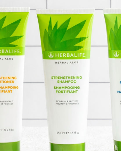 En flaske Herbalife styrkende shampo og balsam på et bord, og i bakgrunnen noen aloe vera-blader som står i et glass vann. 