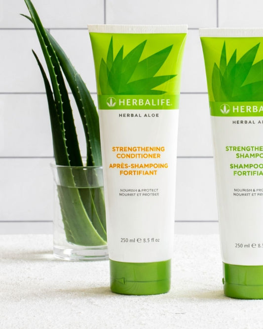 En flaske Herbalife styrkende balsam på et bord sammen med shampo, og i bakgrunnen noen aloe vera-blader som står i et glass vann. 