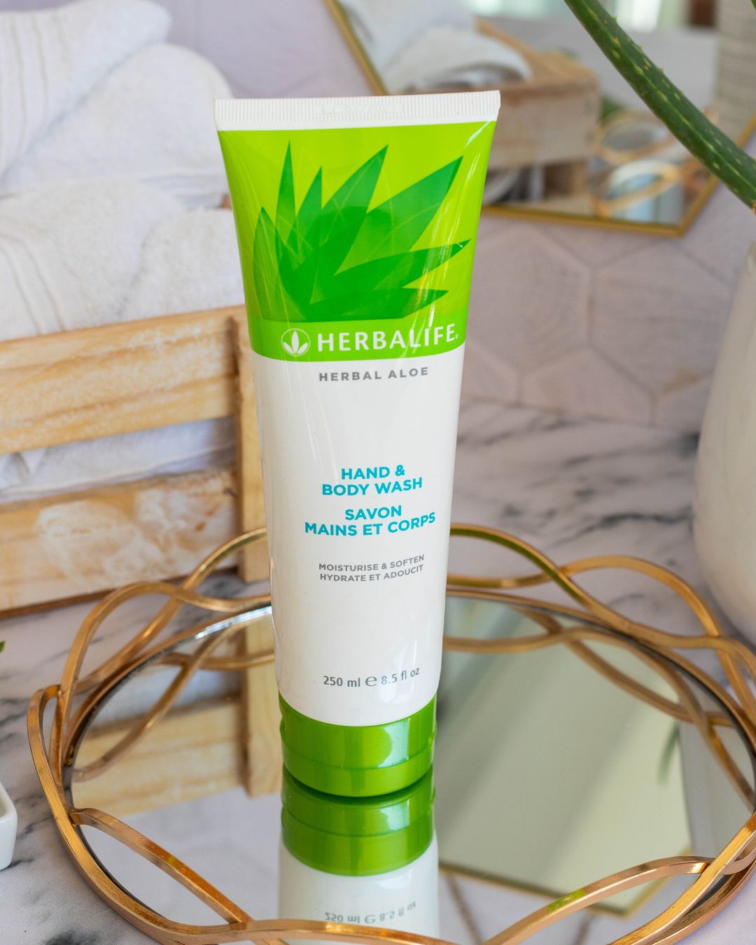 Tube med Herbalife Herbal Aloe Hand & Body Wash på glassfat med gullkant på marmorbord. Aloe vera-plante, oppskårne aloevera-blad og trekasse med hvite håndklær i bakgrunnen.
