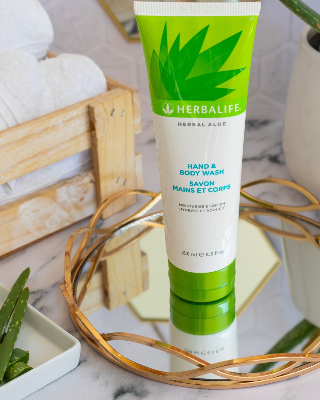 Tube med Herbalife Herbal Aloe Hand & Body Wash på glassfat med gullkant på marmorbord. Aloe vera-plante, oppskårne aloevera-blad og trekasse med hvite håndklær i bakgrunnen.