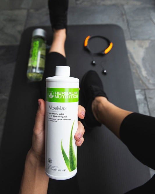 En person i svart treningstøy sitter på ei svart treningsmatte og holder ei flaske Herbalife Herbal Aloe AloeMax urtekonsentrat i hånda.