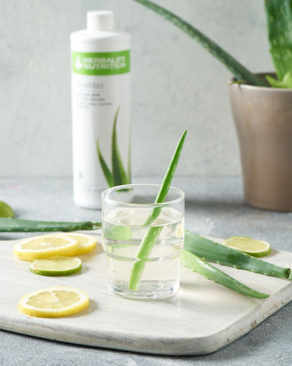 I forgrunnen et glass vann med aloevera-blad i. Aloe vera-blader og skivet sitron og lime ligger på bordet. I bakgrunnen skimtes en flaske Herbalife Herbal Aloe AloeMax urtekonsentrat aloejuice.