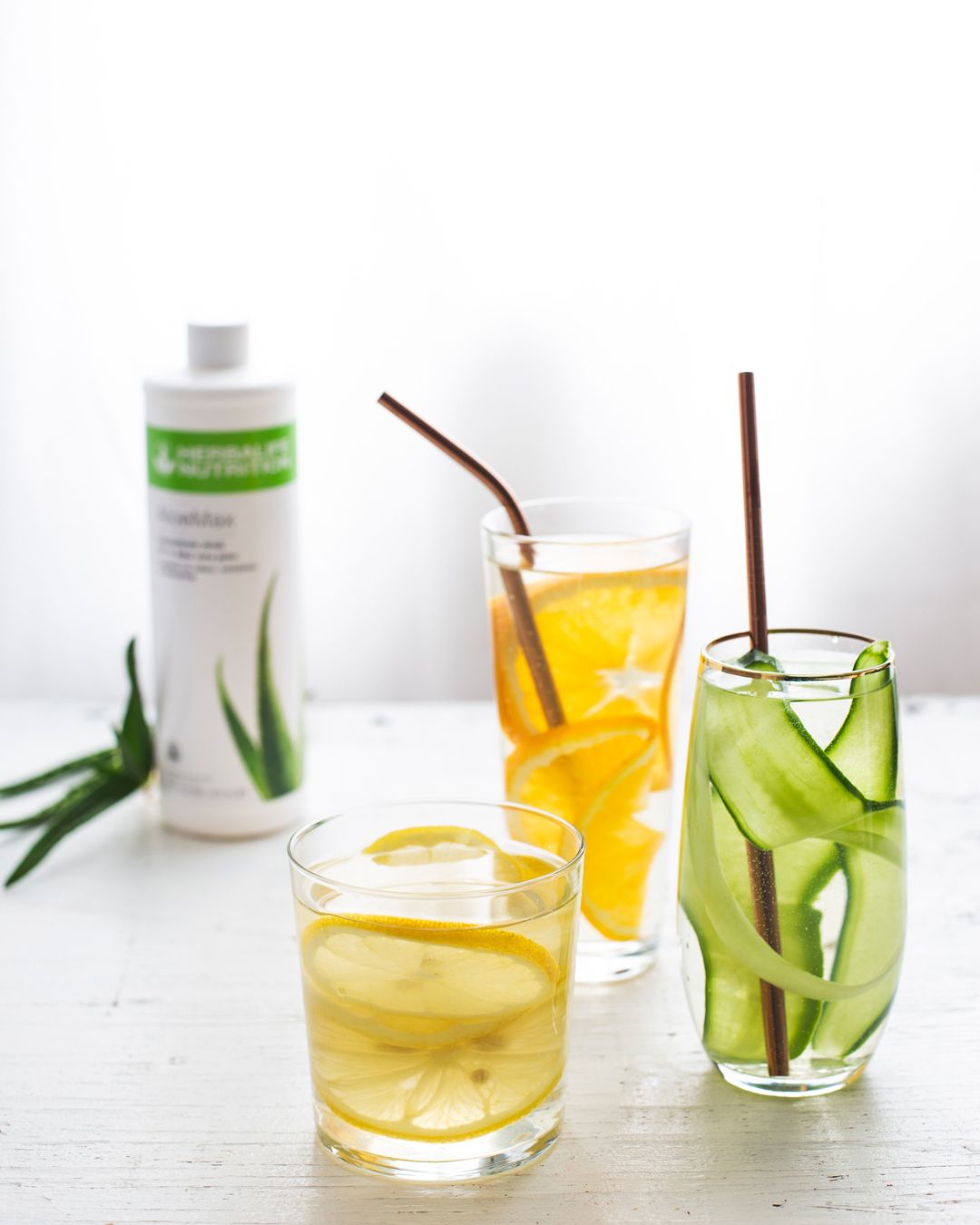 Tre glass vann med aloevera-juice og skivet agurk, appelsin og sitron. I bakgrunnen skimtes ei flaske Herbalife Herbal Aloe AloeMax urtekonsentrat. 