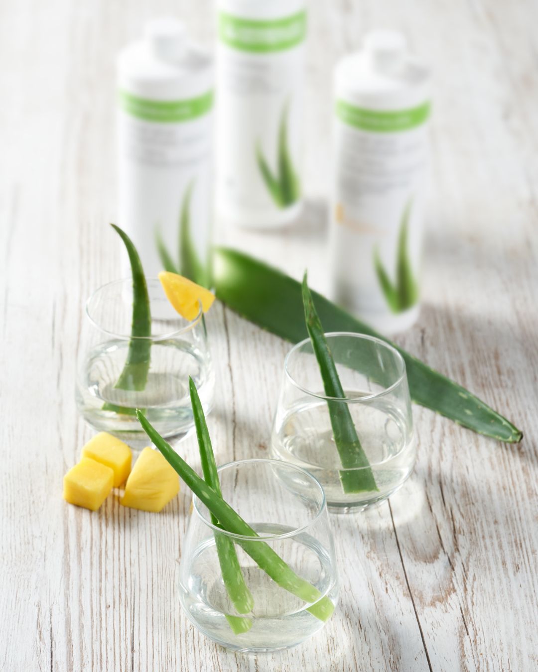 I forgrunnen tre glass vann med aloevera-blad i, det ene glasset pyntet med mango. I bakgrunnen skimtes tre flasker Herbalife Herbal Aloe urtekonsentrat aloejuice. 
