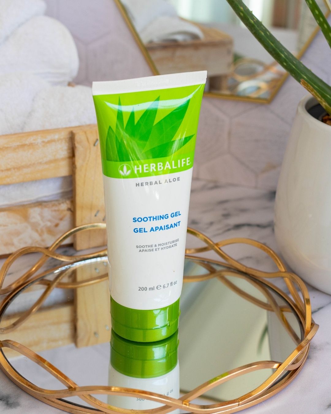Tube med Herbalife Herbal Aloe Soothing Gel på glassfat med gullkant på marmorbord. Aloe vera-plante og oppskårne aloevera-blad og trekasse med hvite håndklær i bakgrunnen.