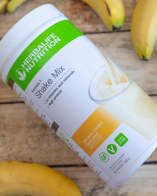 En boks Herbalife F1-shake med banansmak ligger på et trebord sammen med bananer. 
