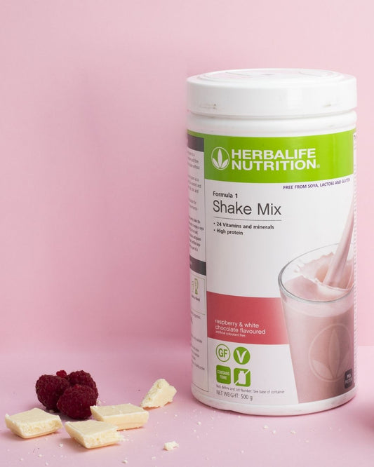 En boks Herbalife Formula 1 soyafri shakemix med Raspberry & White Chocolate-smak og noen biter av hvit sjokolade og hele bringebær ligger på bordet.