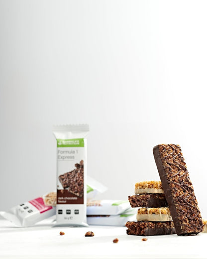 Fire uinnpakkede Herbalife Formula 1 Expressbarer i en stabel, to med tranebær og hvit sjokolade og to med mørk sjokolade. En mørk sjokoladebar lener seg mot stabelen. I den litt uklare bakgrunnen ses flere innpakkede Expressbarer med begge smakene.