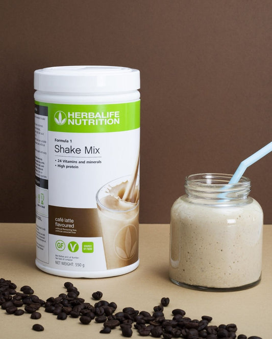En boks Herbalife F1 Caffe latte-shake og en glasstumbler med smoothie og sugerør, i forgrunnen kaffebønner strødd utover et bord. 