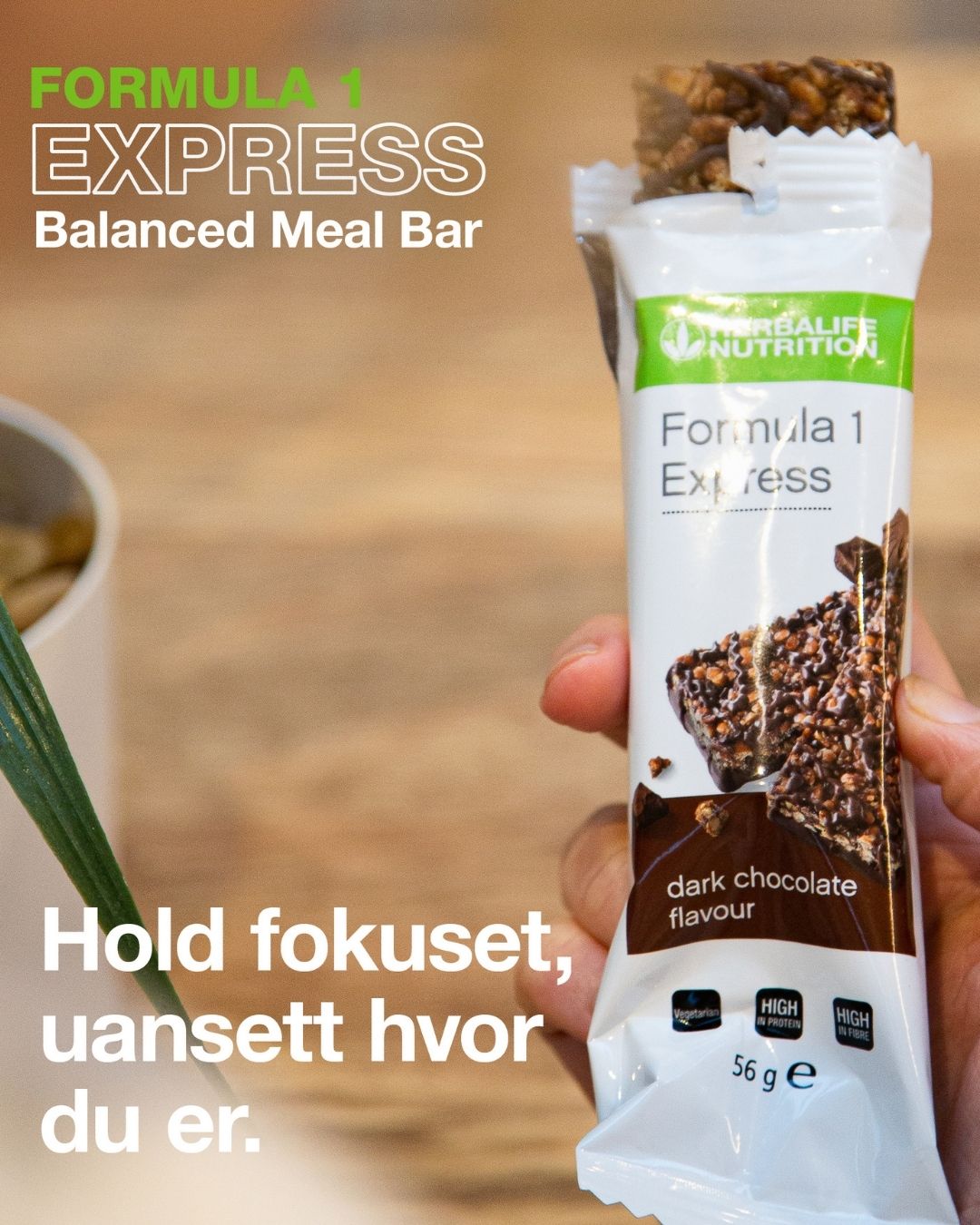 En person holder en åpnet Herbalife Formula 1 mellombar med mørk sjokolade. På bildet står tekstene "Formula 1 Express Balanced Meal Bar" og “Hold fokuset, uansett hvor du er.”.
