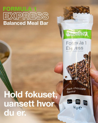 En person holder en åpnet Herbalife Formula 1 mellombar med mørk sjokolade. På bildet står tekstene "Formula 1 Express Balanced Meal Bar" og “Hold fokuset, uansett hvor du er.”.