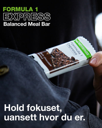 En person i en mørkeblå jakke tar en Herbalife Formula 1 mellombar med mørk sjokolade opp av jakkelomma. På bildet står teksten "Formula 1 Express Balanced Meal Bar".