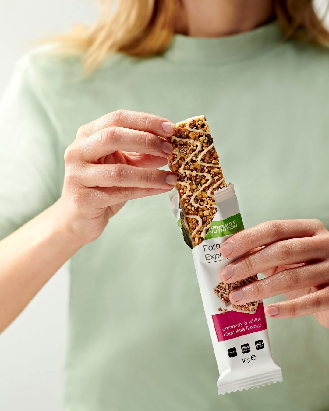 En person holder en Herbalife Formula 1 Express måltidsbar tranebær og hvit sjokolade i hendene og er i ferd med å trekke den ut av papiret.